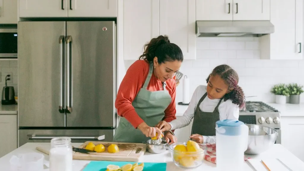 A woman and young girl wearing aprons preparing food together in a modern white kitchen.
