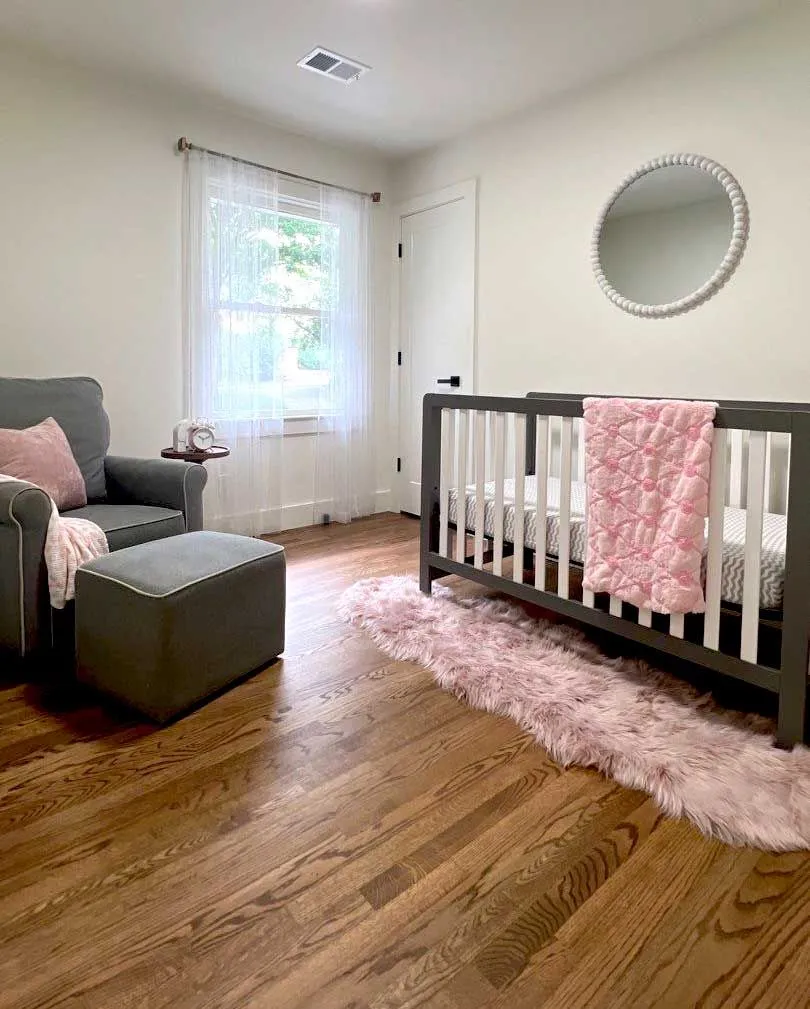 A sunlit baby's room with hardwood floors, a dark gray crib, and a fluffy pink rug.