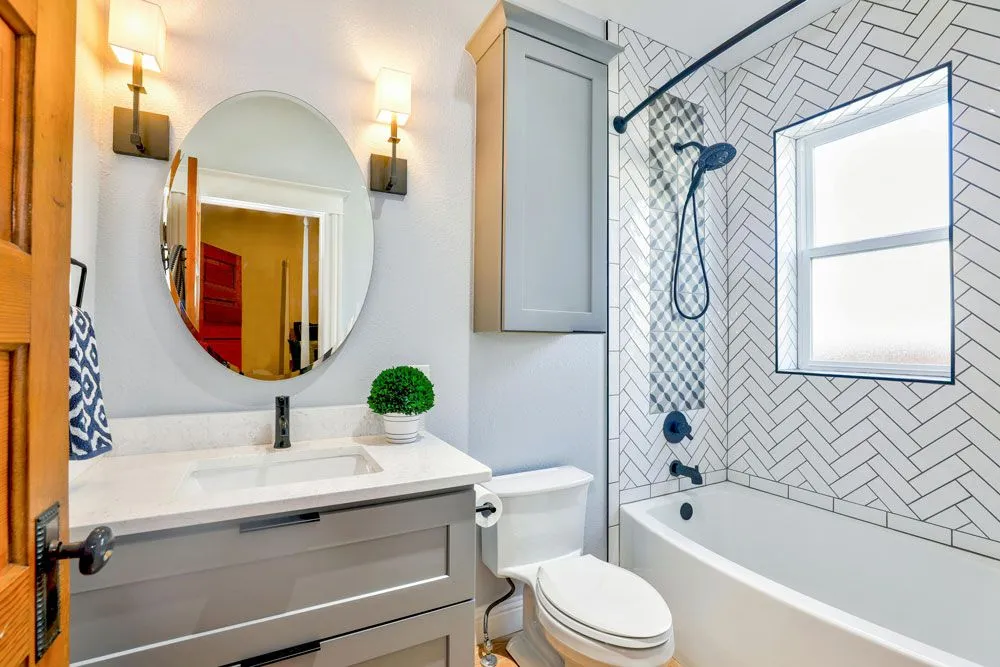 Bright bathroom featuring a gray vanity, round mirror, and herringbone pattern shower tile.